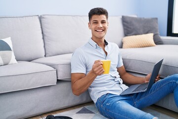 Young hispanic man using laptop and drinking coffee sitting on floor at home