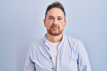 Middle age caucasian man standing over blue background relaxed with serious expression on face. simple and natural looking at the camera.