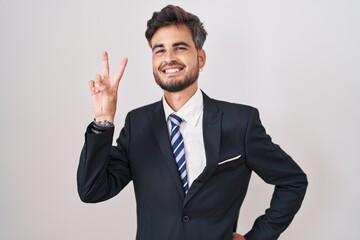 Young hispanic man with tattoos wearing business suit and tie smiling looking to the camera showing fingers doing victory sign. number two.