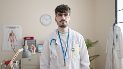 Young hispanic man doctor standing with serious expression at clinic