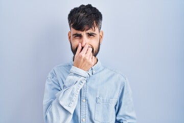 Young hispanic man with beard standing over blue background smelling something stinky and disgusting, intolerable smell, holding breath with fingers on nose. bad smell
