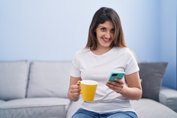 Young hispanic woman drinking coffee and using smartphone at home