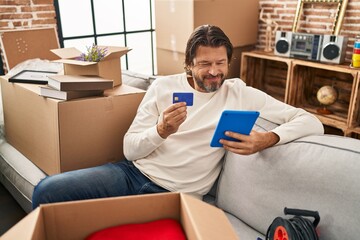 Middle age man using touchpad and credit card sitting on sofa at new home