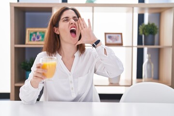 Brunette woman drinking glass of orange juice shouting and screaming loud to side with hand on mouth. communication concept.
