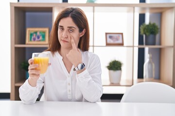 Brunette woman drinking glass of orange juice pointing to the eye watching you gesture, suspicious expression