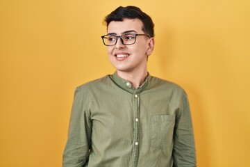Non binary person standing over yellow background looking away to side with smile on face, natural expression. laughing confident.