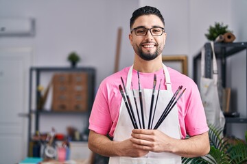 Young arab man artist smiling confident holding paintbrushes at art studio
