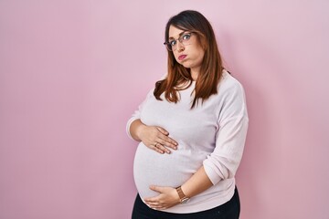 Pregnant woman standing over pink background puffing cheeks with funny face. mouth inflated with air, crazy expression. - obrazy, fototapety, plakaty
