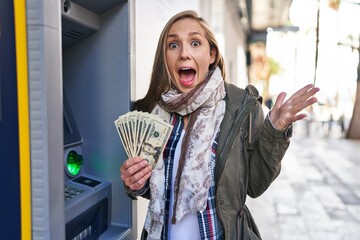 Young blonde woman holding dollars banknotes from atm machine celebrating victory with happy smile...