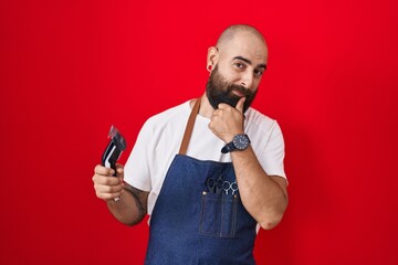 Young hispanic man with beard and tattoos wearing barber apron holding razor looking confident at the camera smiling with crossed arms and hand raised on chin. thinking positive.