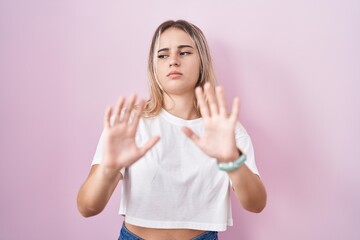 Young blonde woman standing over pink background moving away hands palms showing refusal and denial with afraid and disgusting expression. stop and forbidden.