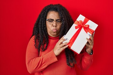 Plus size hispanic woman holding gift skeptic and nervous, frowning upset because of problem. negative person.