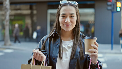 Young beautiful hispanic woman smiling confident holding shopping bags and coffee at street