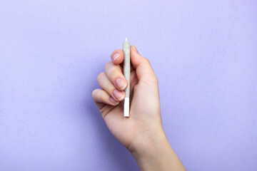 A woman's hand with natural nails holds a joint with medical marijuana on a light purple background.  copy space