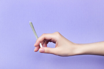 A woman's hand with natural nails holds a joint with medical marijuana on a light purple background.  copy space