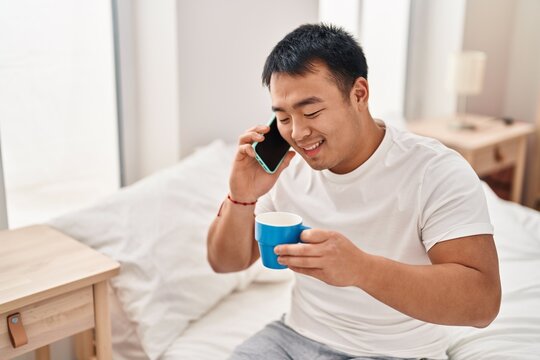 Young chinese man talking on the smartphone drinking coffee sitting on bed at bedroom