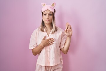 Blonde caucasian woman wearing sleep mask and pajama swearing with hand on chest and open palm,...