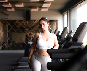 Fototapeta na wymiar Sexy Indian Girl in a sports bra in a Gym standing next to a Treadmill. Confident Indian Fitness Model.