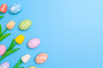 Happy Easter holiday greeting card design concept. Colorful Easter Eggs and spring flowers on blue background. Flat lay, top view, copy space.