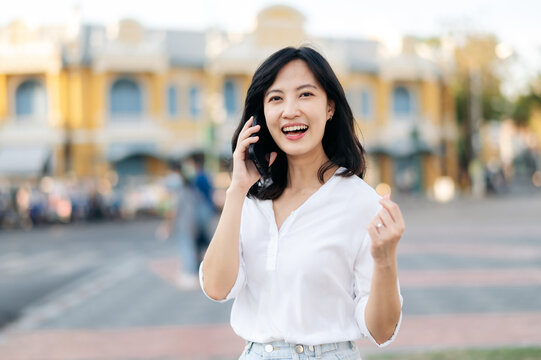 Portrait beautiful young asian woman receiving good news on mobile phone and arms raised feeling cheerful ,happiness. around outdoor street view in a summer day