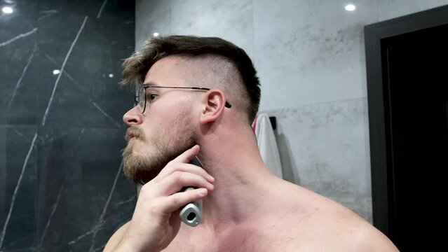Beautiful muscular man shawing his beard with electric trimmer. Wearing dioptrical round glasses. In a marbel stone bathroom. Grooming and looking in to the mirror. Brown hair blue eyes.