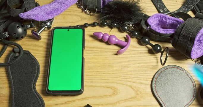 Man places several anal stimulators on table for comparison. Smartphone with green screen. Concept of online service, store, shopping, sex shop, information search.