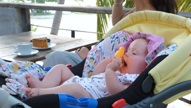 Cute baby girl playing with toy in stroller while mother drinking coffee and eating cake on a terrace of cafe or restaurant in tropics. Summer vacation and travel concept