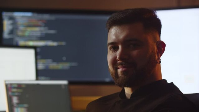 A well-groomed young man smiles at the camera. Close-up of a smiling male developer sitting in his office in front of monitors with source code.
