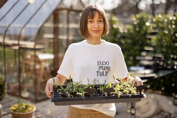 Portrait of young female gardener with a seedling tray in beautiful garden during early spring with...