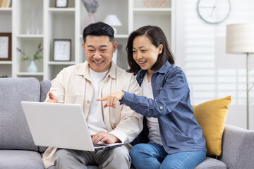 Happy asian family, couple in love man and woman together at home sitting on sofa happy in living room choosing products in online store using laptop spending time together.