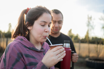 Smiling friends drinking yerba mate using a thermo with hot water in the countryside at sunset....