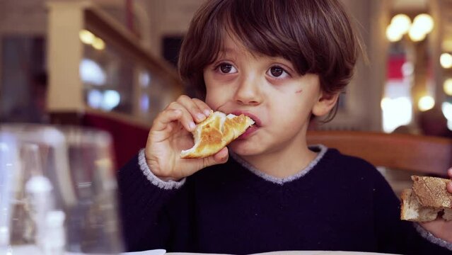 Child eating bread sitting at restaurant. One little boy eats piece of carb food croissant