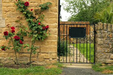Fototapeta na wymiar Black metal gate into garden next to stone English cottage with red rose bush on the wall, on a summer day .
