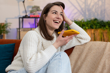 Portrait of happy excited young woman enjoying mobile smartphone loudspeaker talking good news, laughing at home room apartment. Pretty girl making phone call conversation with friends sitting on sofa