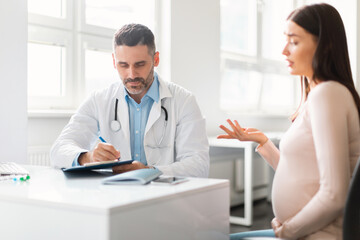 Young expecting woman visiting doctor man at private clinic, therapist giving recommendations to pregnant lady