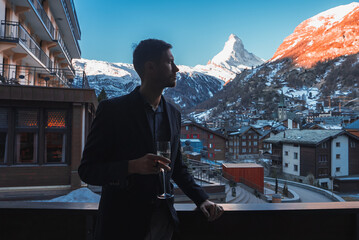 Cheerful man in a suit enjoying the view of the Matterhorn peak in Zermatt from a balcony. He is standing and relaxing. Focus on his back. Luxury winter resort concept.