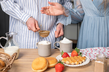 Obraz na płótnie Canvas A mans and a woman's hands in light blue clothes in the background. A man ads cinnamon in a gray cup with caffe latte. The crispy waffle, red strawberries on a white plate and donuts.