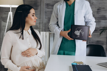 Doctor showing a pregnant girl picture ultrasound. She came to the doctor in the clinic. It's a gynecologist. He is dressed in a white robe
