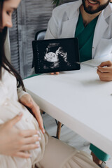 Doctor showing a pregnant girl picture ultrasound. She came to the doctor in the clinic. It's a gynecologist. He is dressed in a white robe