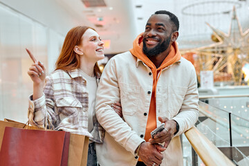 smiling cheerful red-haired girl showing something in the shop holding her boyfriend arm, asking him to buy clothes, goods, present for herself