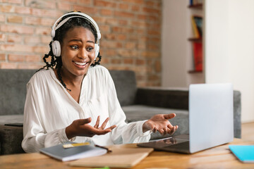 Video call. Emotional black woman in headphones talking and gesturing at laptop webcamera, sitting...