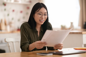 Happy asian woman sitting at kitchen desk, holding papers