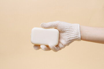 Female hand in body scrub glove holding natural soap on beige isolated background. Concept of skin care, beauty products and spa.