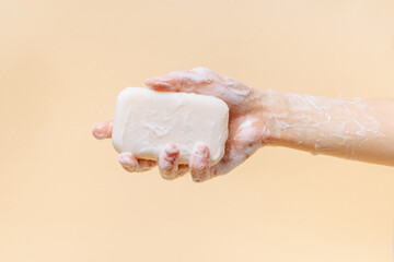 Female hand covered with foam holding natural soap on beige isolated background. The concept of cleanliness, self-care and disinfection