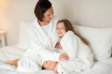 Obraz na płótnie Canvas Cheerful young caucasian female hugging little girl in bathrobe, sitting on white soft bed, enjoy weekend