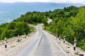 Rural tarmac winding downhill road covered with loose stones and gravel from Khvamli mountain peak down to Tskhenistsqali river valley with lush green trees around and mountain landscape.