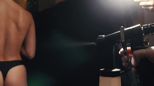 Cosmetologist using airbrush for spray tan apply to young woman body in beauty salon. 