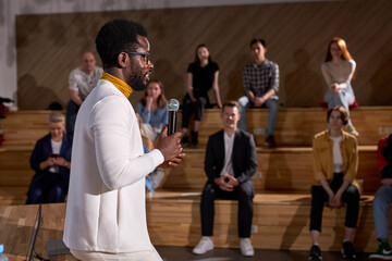 Side View Of Nice African American Teacher In White Clothing Lecturing Presenting Speech Holding Microphone In Hands, Discussing Important Theme With Students, Group Of People In The Background