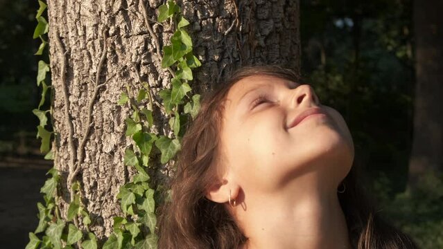 Healthy child time among trees. A view of healthy girl stay by the tree trunk and enjoy the clean forest air. A concept of eco friendly movement during child life.