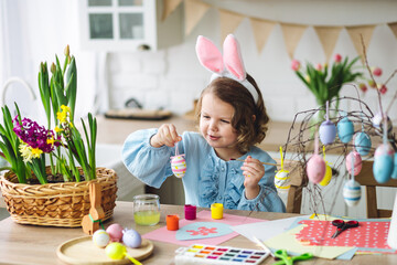 Portrait of cute pretty little girl in blue dress and funny rabbit ears making craft, diy eggs for easter holiday home decoration. Spring colours, bright flowers. Kid's domestic life, hobby, enjoyment